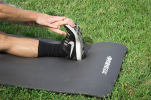 picture of a caucasian male stretching their leg on a black mat. Pictured on a grass field. 
